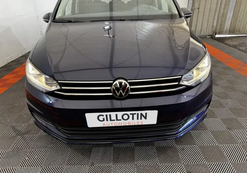 Vue avant rapprochée d’un Volkswagen Touran bleu 2024 avec calandre chromée et phares allumés.