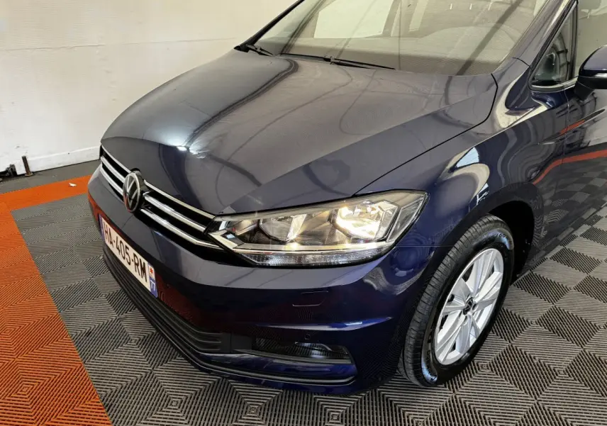 Vue 3/4 avant droit d’un Volkswagen Touran bleu 2024 avec phares allumés et jantes argentées.
