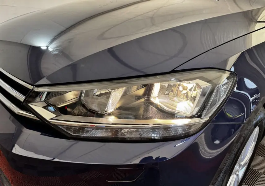 Gros plan sur le phare avant droit d’un Volkswagen Touran bleu 2024, mettant en valeur la calandre chromée et la carrosserie brillante.