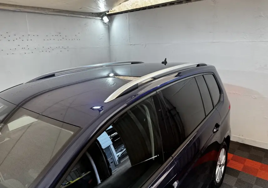 Vue latérale droite du Volkswagen Touran bleu 2024 avec barres de toit argentées et vitres teintées dans un garage.