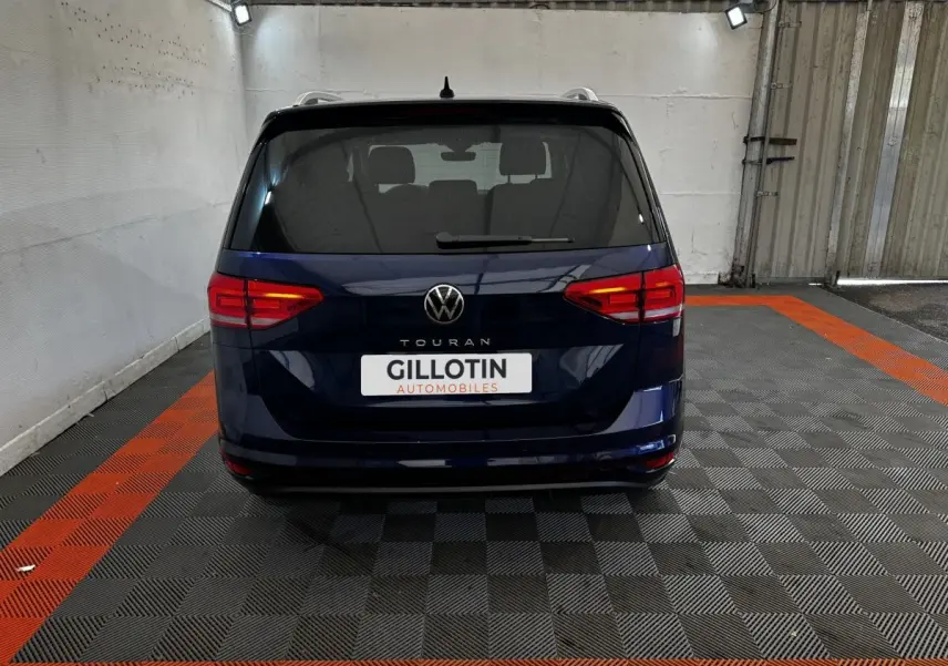 Vue arrière d'un Volkswagen Touran bleu 2024 garé sur un sol à damiers noir et orange dans un garage.