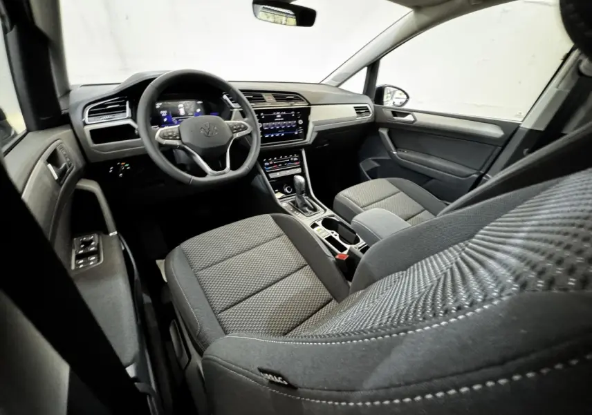 Vue intérieure côté conducteur du Volkswagen Touran 2024, montrant le tableau de bord et les sièges en tissu gris.