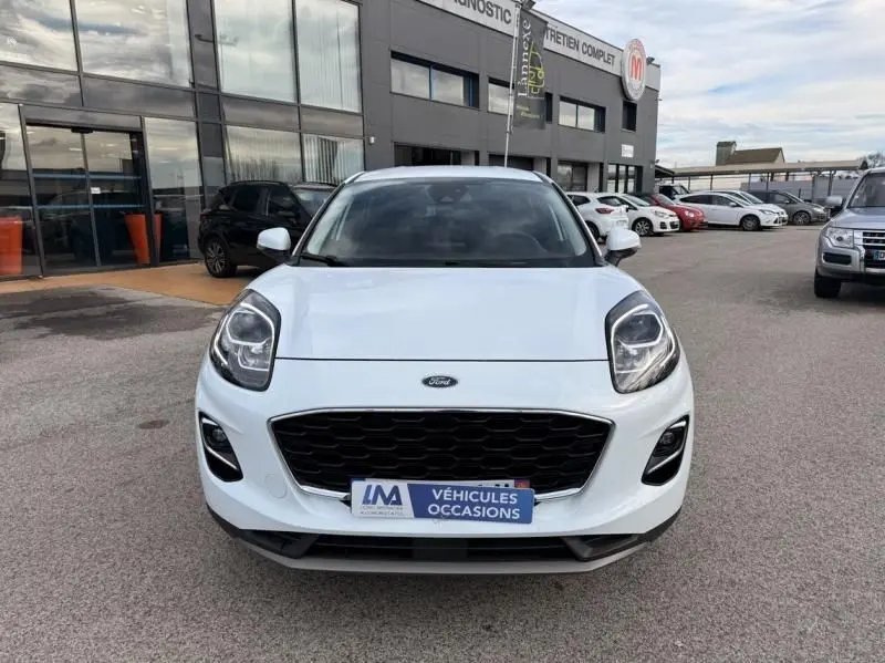 Vue frontale d'un Ford Puma blanc 2026 avec calandre noire et plaque "Véhicules Occasions" sur un parking extérieur.