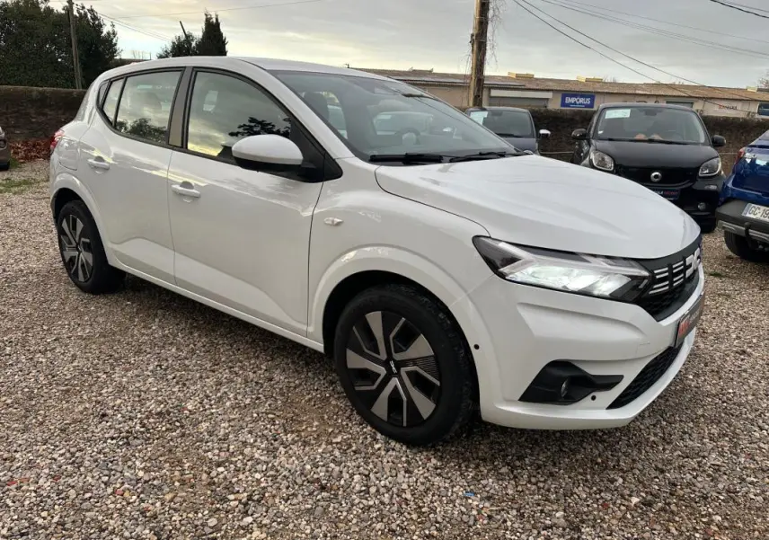 Vue 3/4 avant droit d'une Dacia Sandero TCe 90 Expression blanche avec jantes noires et phares allumés.