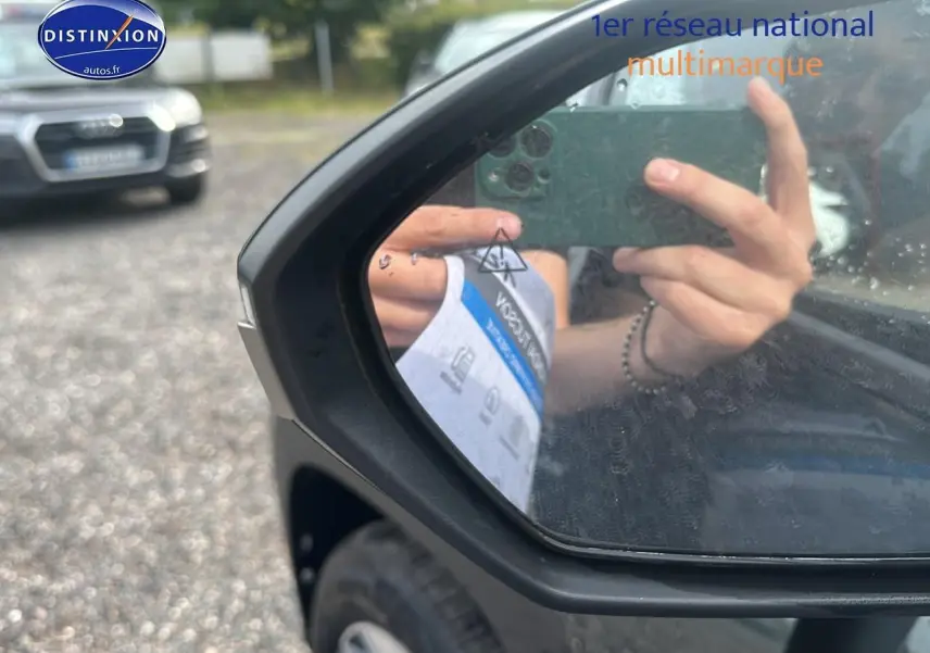 Rétroviseur côté gauche d'un Hyundai Tucson vert cyprès 2025, avec reflet d'une main tenant un smartphone.