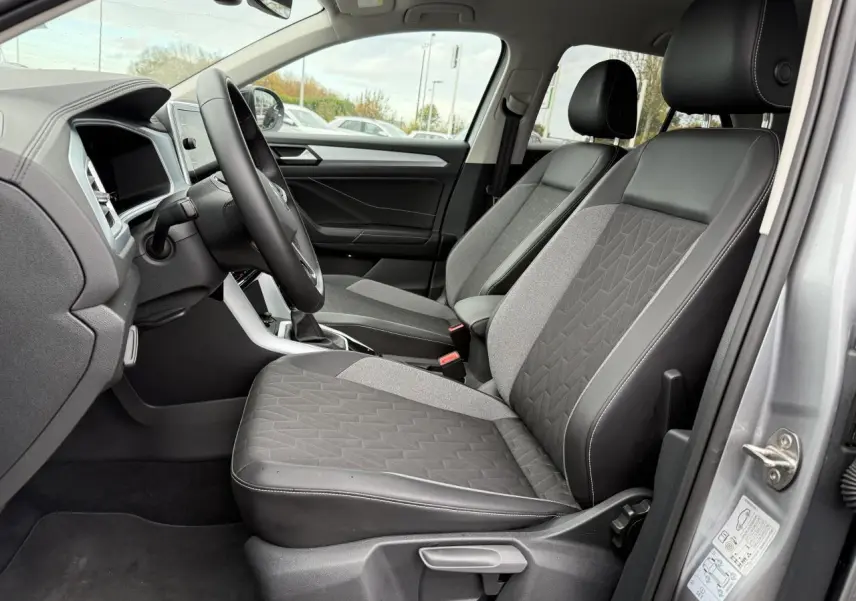 Vue intérieure côté conducteur du Volkswagen T-Roc 2025 argent pyrite, sièges avant en tissu et cuir noir avec surpiqûres blanches.