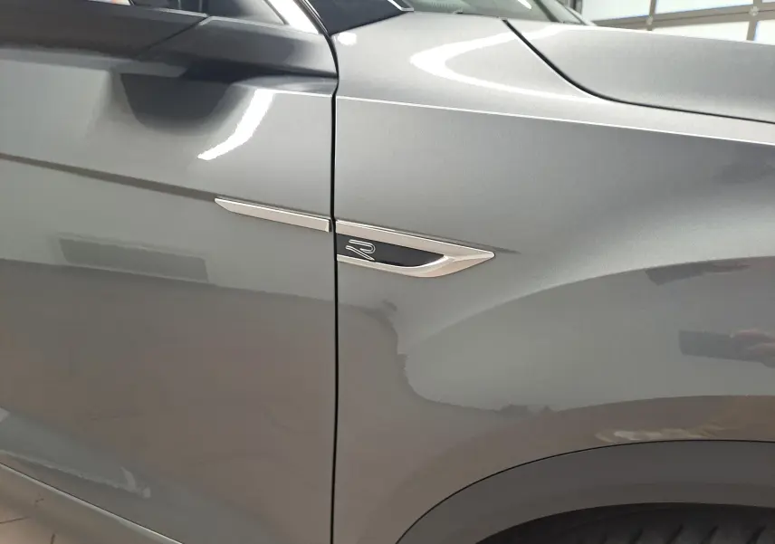 Gros plan sur l'aile avant gauche du Volkswagen T-Roc gris Indium, avec l'emblème R-Line chromé visible.