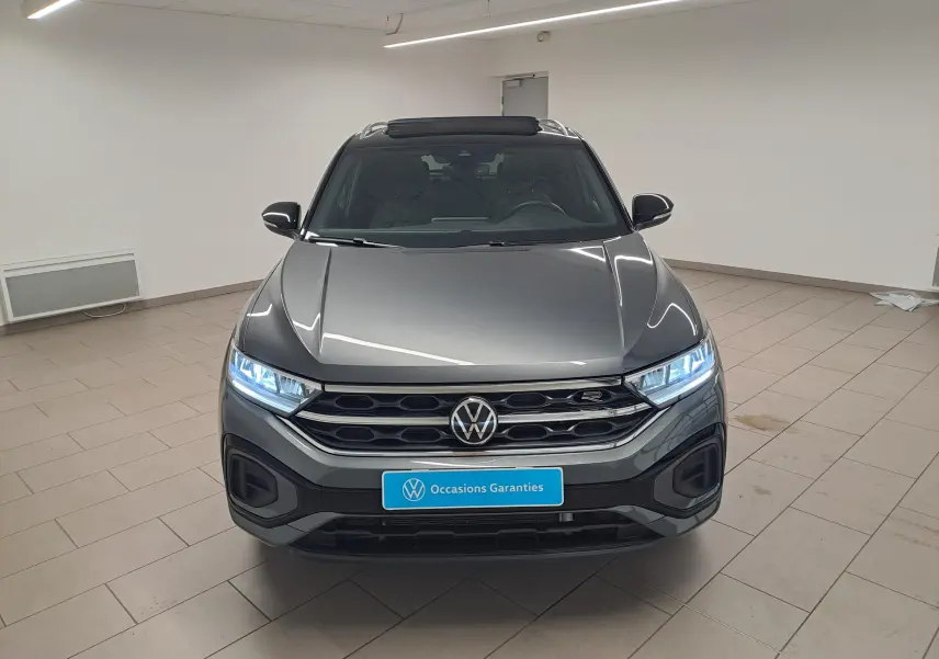 Vue frontale d'un Volkswagen T-Roc 2025 gris Indium avec toit noir et phares LED allumés en intérieur showroom.