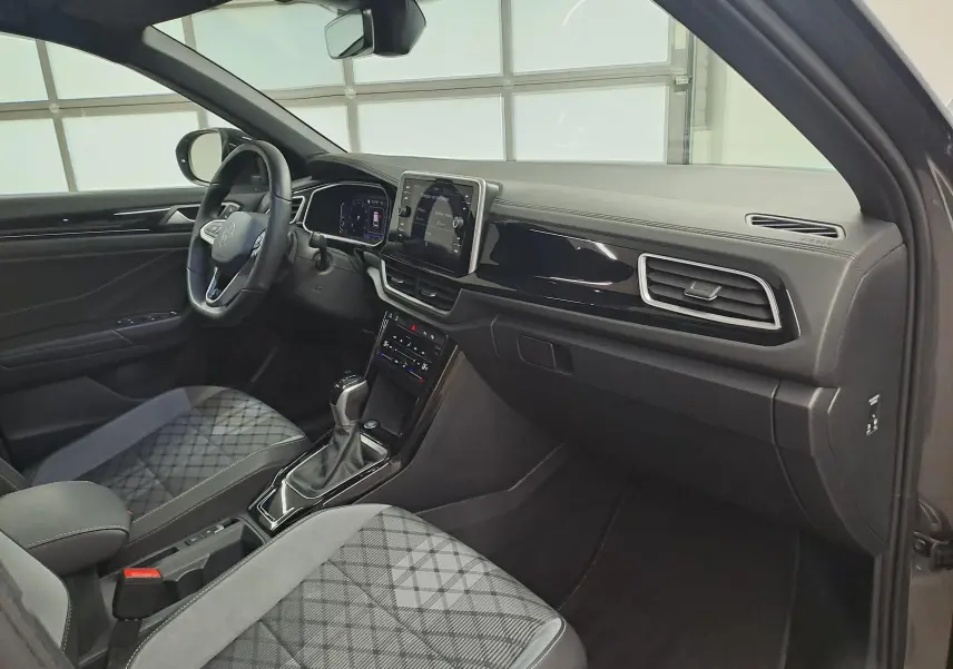 Vue intérieure côté passager du Volkswagen T-Roc 2025, tableau de bord noir avec sièges tissu gris à motif géométrique.