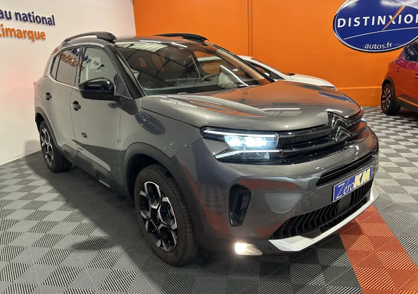Vue 3/4 avant d'un Citroën C5 Aircross gris platinium avec phares allumés dans un showroom orange.