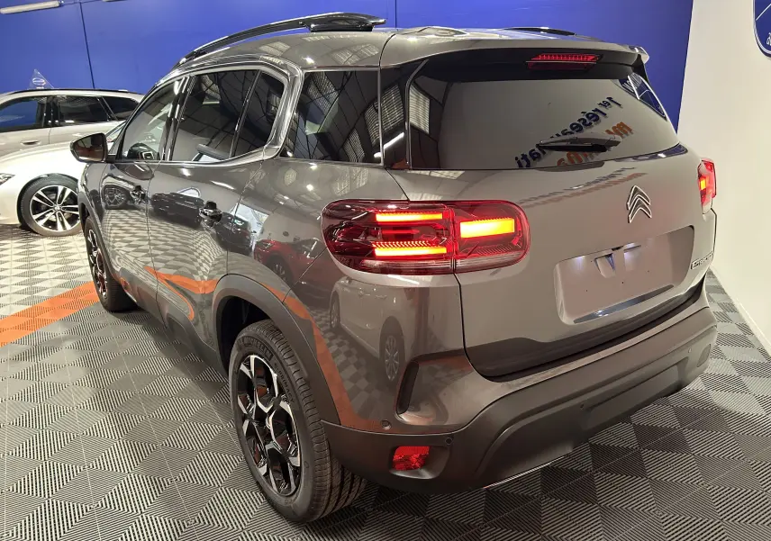 Vue 3/4 arrière droite d'un Citroën C5 Aircross gris platinium avec feux arrière allumés en intérieur showroom.