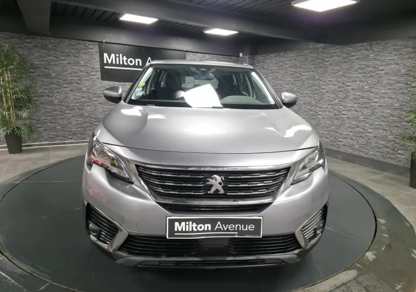 Vue avant d'un Peugeot 5008 gris métallisé exposé en showroom avec calandre chromée et phares allumés.