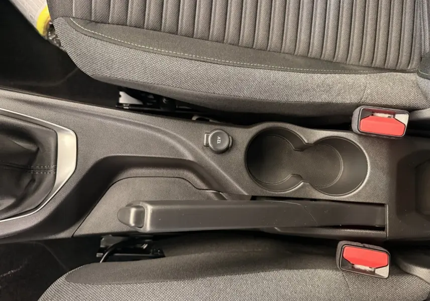 Vue plongeante sur la console centrale noire de la Peugeot 208 blanche, avec frein à main, prise 12V et porte-gobelets.