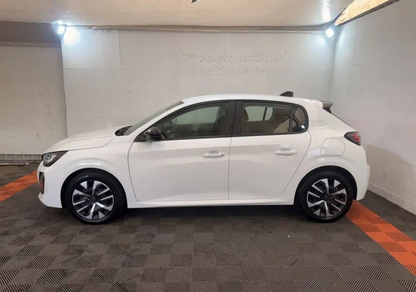 Profil côté gauche d'une Peugeot 208 blanche 2025 avec jantes noires et grises dans un garage éclairé.