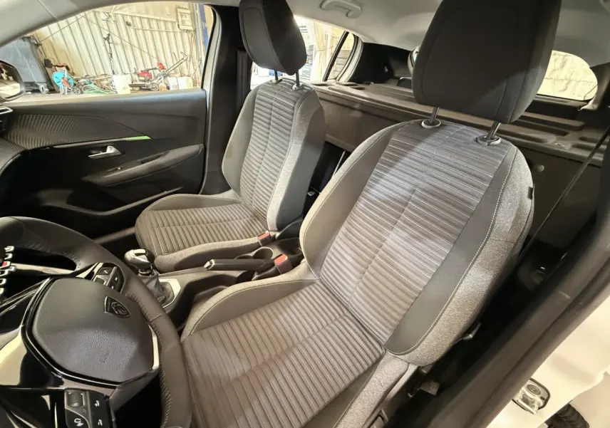 Vue intérieure côté conducteur de la Peugeot 208 blanche 2025, sièges tissu gris et volant cuir multifonctions.