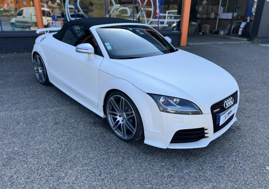 Audi TT RS Roadster blanc vue 3/4 avant avec capote noire et jantes sport grises sur parking.