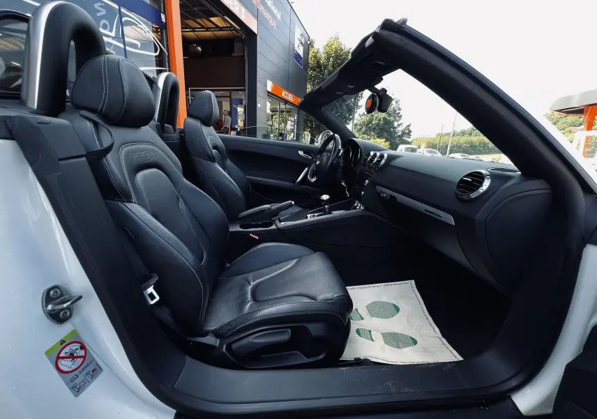 Vue intérieure côté conducteur de l'Audi TT RS Roadster blanc, mettant en valeur les sièges baquet cuir noir et le tableau de bord.