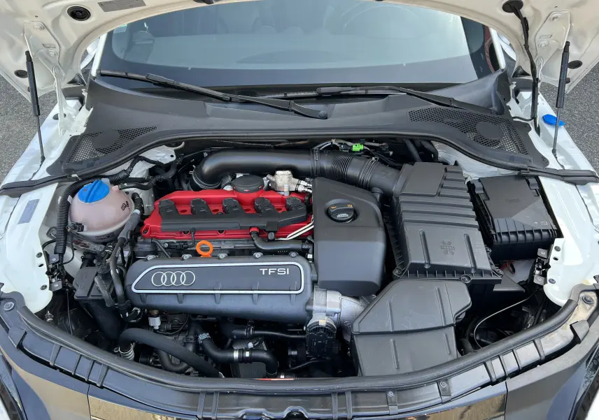 Moteur TFSI rouge et noir visible sous le capot ouvert d'un Audi TT RS Roadster blanc, vue de face.