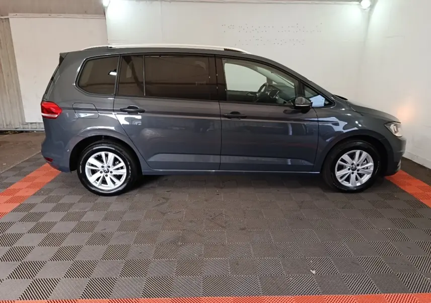Vue de profil côté gauche d'un Volkswagen Touran gris clair 2024 dans un garage avec sol à damiers noir et orange.