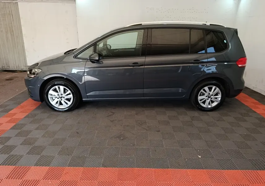 Vue latérale droite d'un Volkswagen Touran gris clair 2024, stationné sur un sol à damiers gris et rouge.