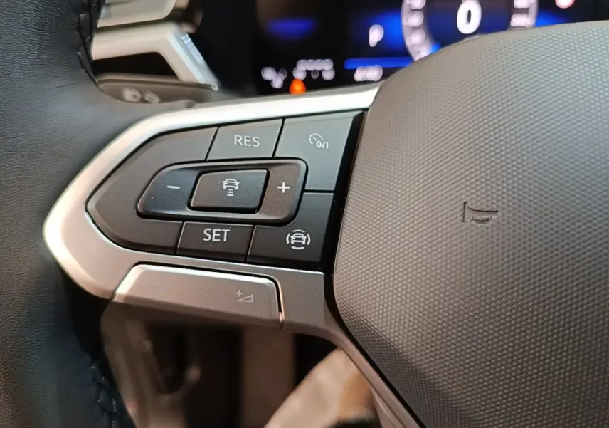 Gros plan sur les commandes à gauche du volant du Volkswagen Touran 2024, boutons noirs et argentés avec affichage numérique flou en arrière-plan.