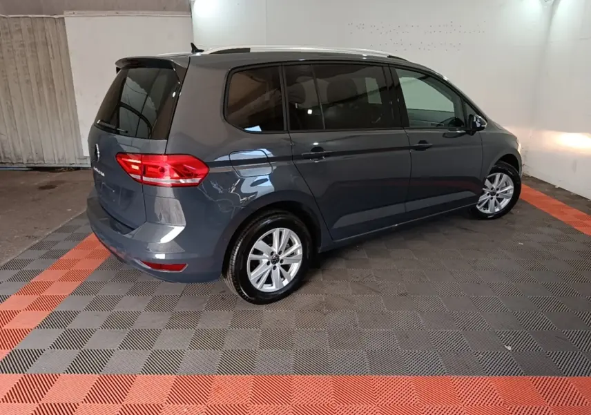 Vue 3/4 arrière droite d'un Volkswagen Touran gris clair 2024 stationné en intérieur sur sol à damiers noir et rouge.