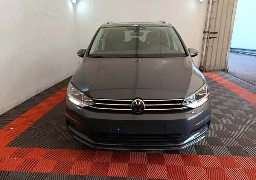 Vue avant d'un Volkswagen Touran gris clair 2024 avec phares allumés dans un garage fermé.