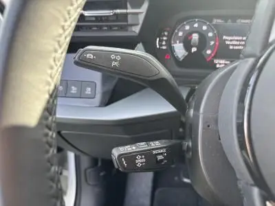 Gros plan sur les commandes au volant d'une Audi A3 Sportback 2024, avec tableau de bord numérique en arrière-plan.