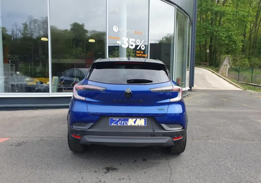 Vue arrière d’un Renault Captur bleu Iron 2026 avec feux LED et plaque Zero KM devant un bâtiment vitré.