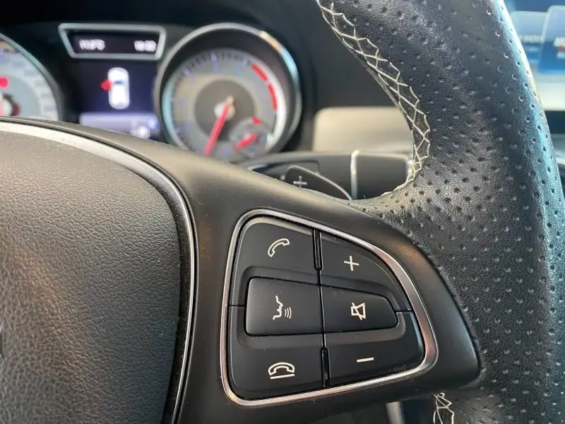 Gros plan sur les commandes du volant en cuir perforé d'une Mercedes GLA 200 Sensation gris montagne, tableau de bord flou en arrière-plan.