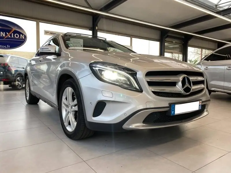 Mercedes GLA 200 Sensation gris montagne en 3/4 avant droit, phares allumés dans un showroom lumineux.