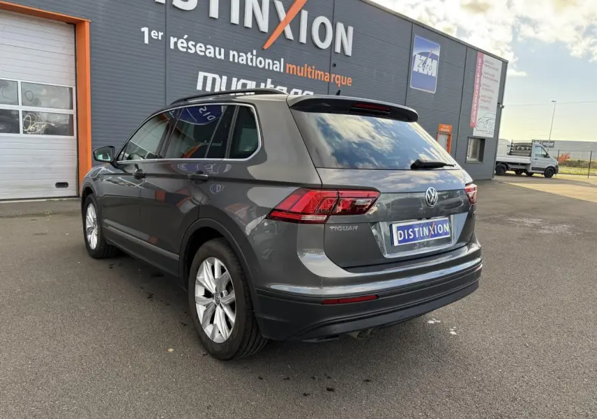 Vue 3/4 arrière droite d'un Volkswagen Tiguan gris Indium avec vitres arrière surteintées, garé devant un bâtiment commercial.
