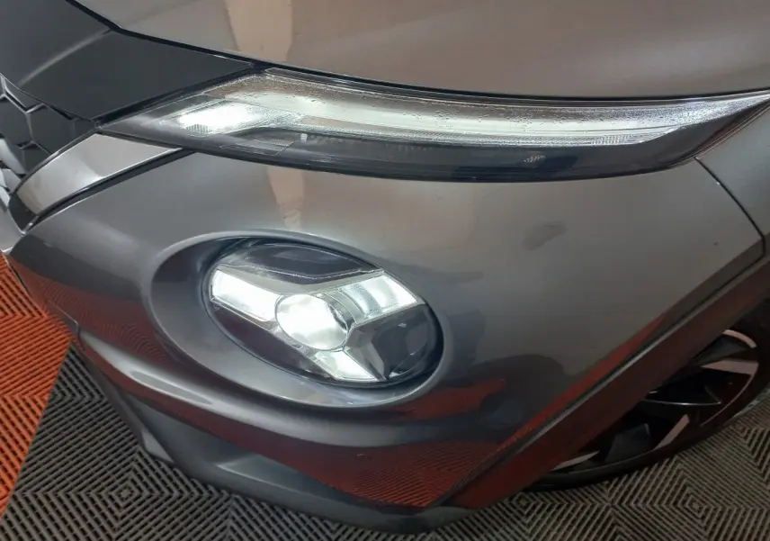 Gros plan sur le phare avant droit allumé d'un Nissan Juke Hybrid 143 Business+ gris foncé, avec partie de la calandre visible.