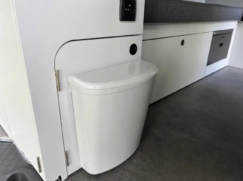 Détail intérieur du fourgon Renault Trafic aménagé, montrant une poubelle blanche intégrée près d'un meuble blanc.