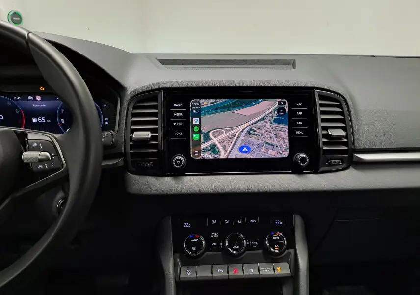 Vue intérieure centrée sur le tableau de bord et l'écran tactile du Skoda Karoq 1.5 TSI blanc Lune, avec commandes climatisation visibles.