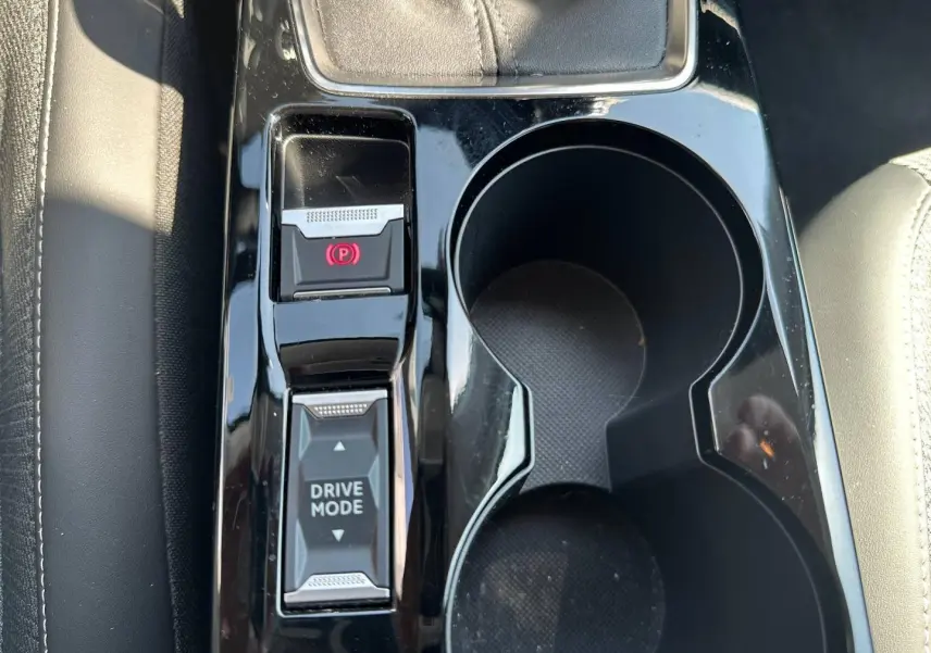 Gros plan sur la console centrale noire brillante de la Peugeot 208 1.2 100ch, avec commandes frein à main électrique et drive mode.