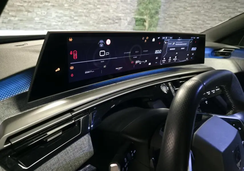 Vue intérieure du tableau de bord numérique panoramique du Peugeot 3008 hybride 2025, avec volant noir et finitions tissu gris.