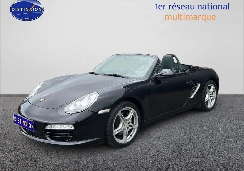 Vue 3/4 avant droite d'une Porsche Boxster 987 noire cabriolet avec jantes alu 17 pouces sur sol gris.