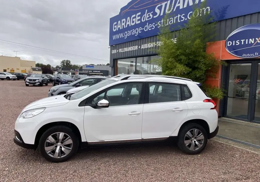 Profil côté gauche d'un Peugeot 2008 blanc 2015 garé devant un garage, avec jantes alliage et rétroviseurs chromés.