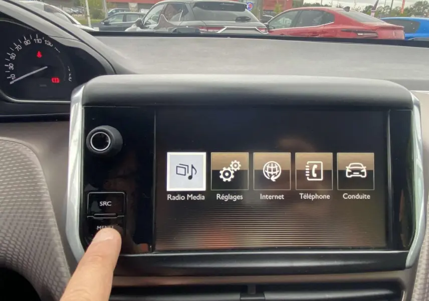 Vue intérieure du tableau de bord du Peugeot 2008 blanc de 2015, avec écran tactile et main appuyant sur le bouton menu.