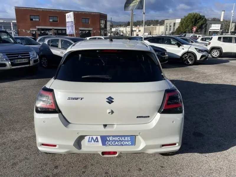 Vue arrière d'une Suzuki Swift blanche 2025 avec badges Swift et Hybrid, stationnée sur un parking extérieur.