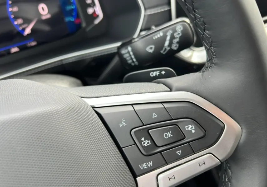 Gros plan sur les commandes multifonctions du volant cuir du Volkswagen T-cross gris argent, avec tableau de bord digital en arrière-plan.