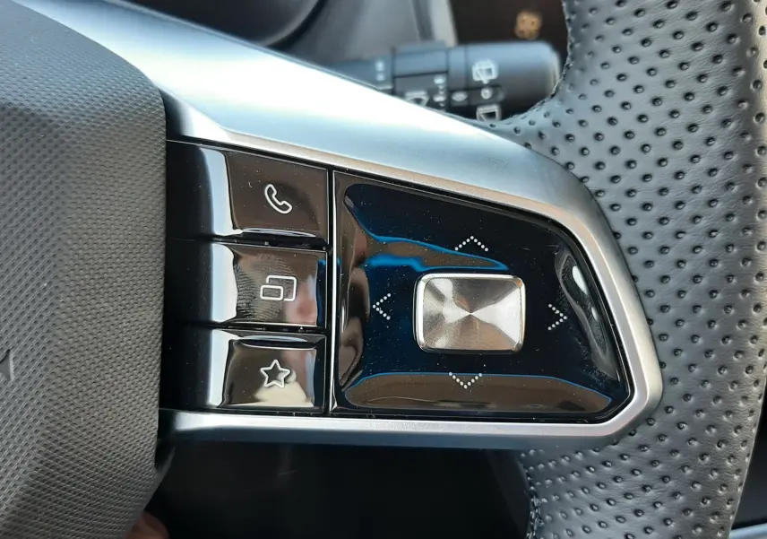 Gros plan sur les commandes gauche du volant perforé du MG ZS 1.5 Hybrid+ 2026, finition noire brillante et métal brossé.