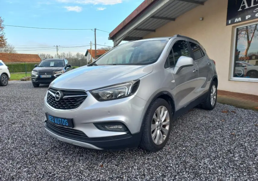 Opel Mokka X gris vue 3/4 avant droit, avec calandre noire et jantes alliage, stationné sur gravier devant un bâtiment.
