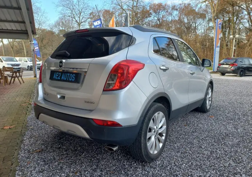 Opel Mokka X gris 2017 vue 3/4 arrière droit, avec feux rouges et jantes alliage visibles, stationné sur gravier.