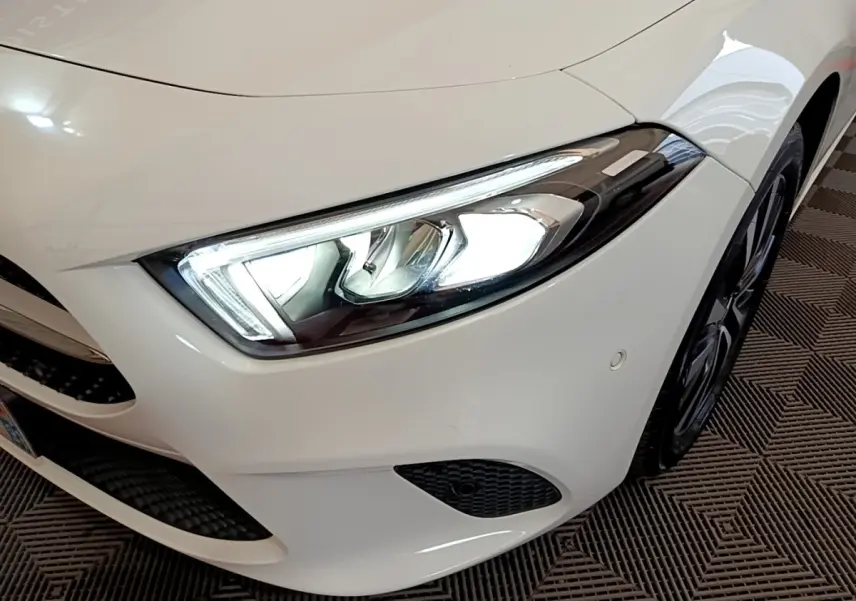 Gros plan sur l'avant droit blanc d'une Mercedes Classe A Berline avec phare LED allumé et jante noire visible.
