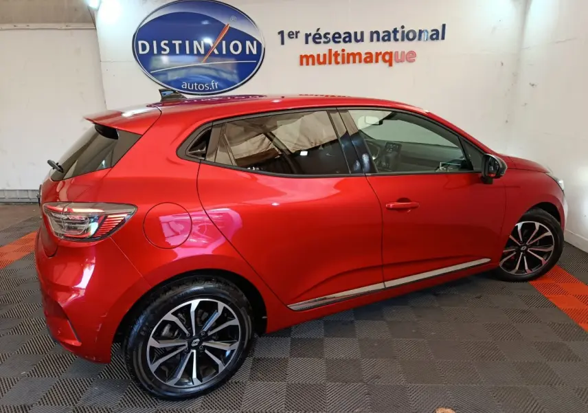 Profil droit d'une Renault Clio Techno TCe 90 rouge 2024 avec jantes alliage et vitres teintées en intérieur showroom.