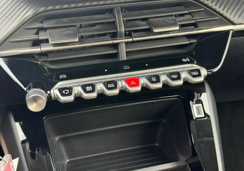 Gros plan sur la console centrale de la Peugeot 208 blanche, montrant les commandes climatisation et bouton rouge de warning.
