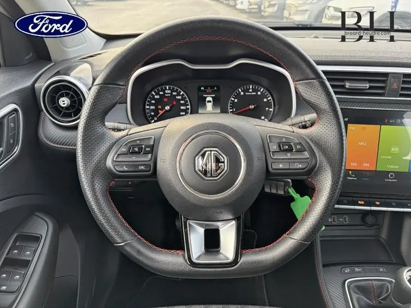 Vue intérieure centrée sur le volant cuir noir à surpiqûres rouges du MG ZS Cosmic Silver avec tableau de bord et écran tactile.