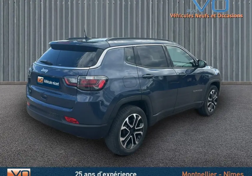 Jeep Compass bleu 3/4 arrière droit, avec barres de toit et jantes 18 pouces visibles, stationné devant un mur métallique.